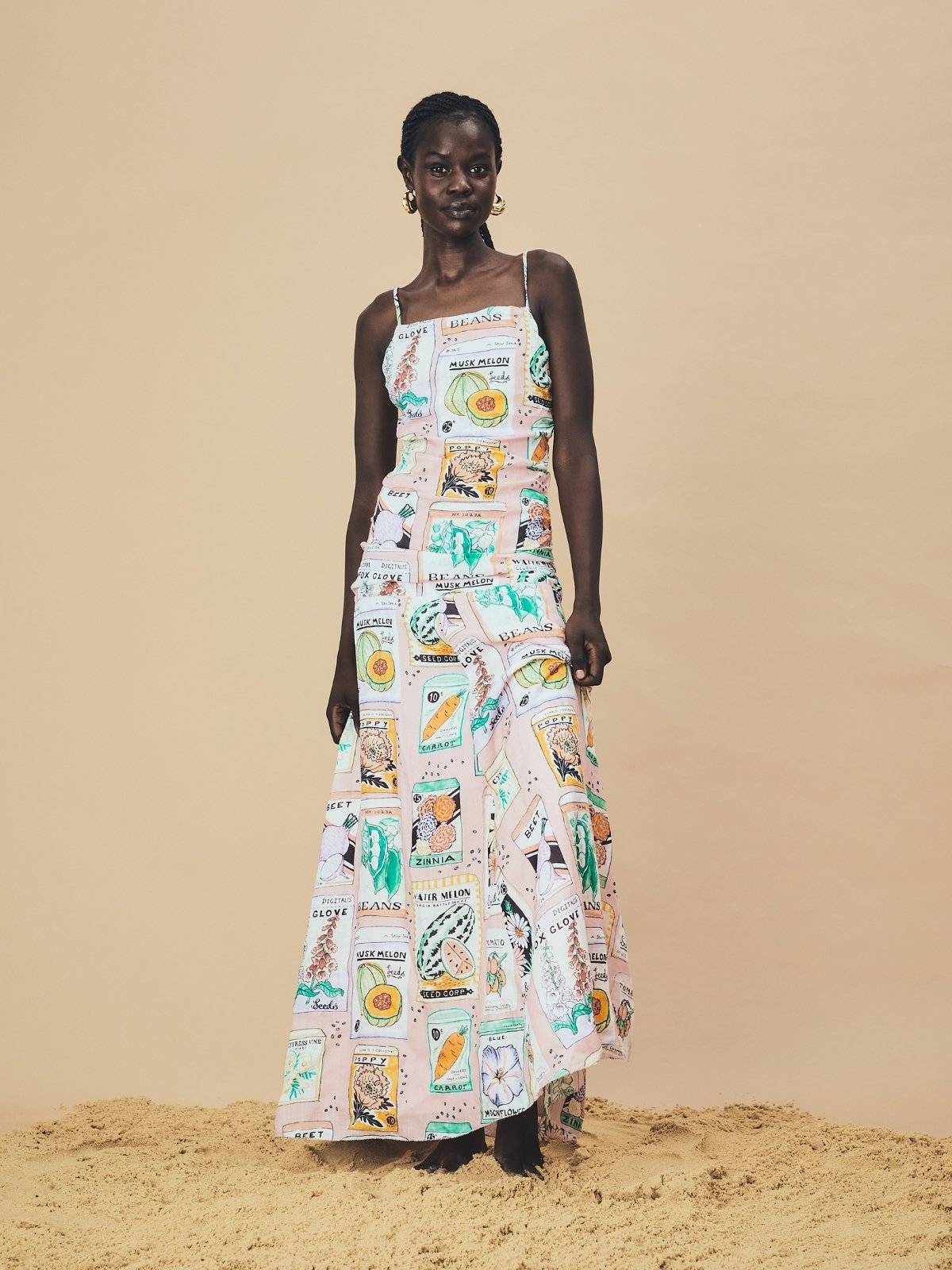 A woman stands barefoot on sand wearing a full-length sleeveless dress made from 100% ramie, featuring a playful vintage seed-packet print in pastel tones. The dress has thin straps, a fitted bodice, and a flowing skirt, and the model poses confidently against a neutral background.