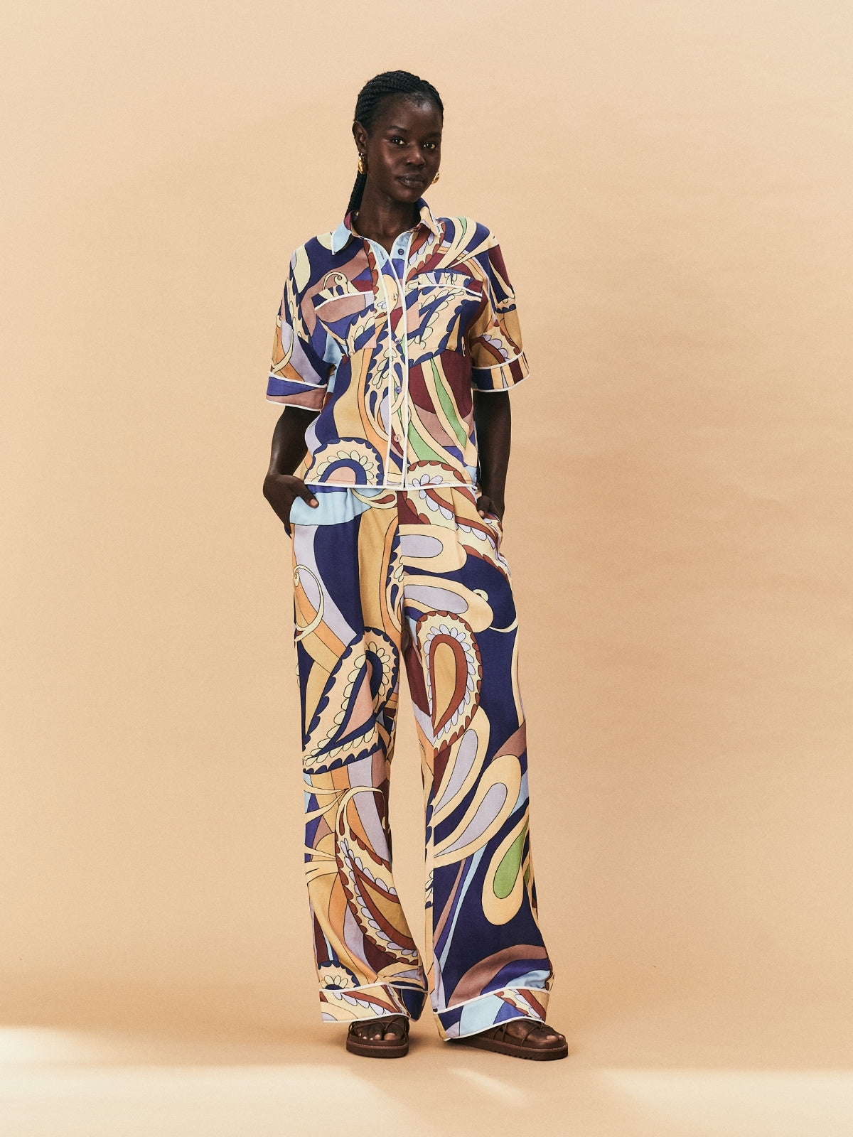 A woman wearing a colourful patterned La Maise Dijon pants with a shirt outfit on a beige background.
