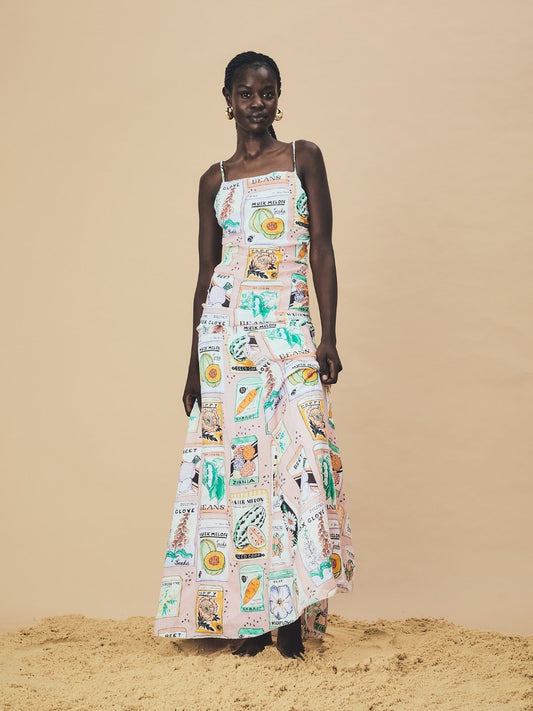 A woman stands barefoot on sand wearing a full-length sleeveless dress made from 100% ramie, featuring a playful vintage seed-packet print in pastel tones. The dress has thin straps, a fitted bodice, and a flowing skirt, and the model poses confidently against a neutral background.