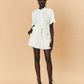 A young woman wearing a white La Maise Lillie short with gold accents on a beige background.