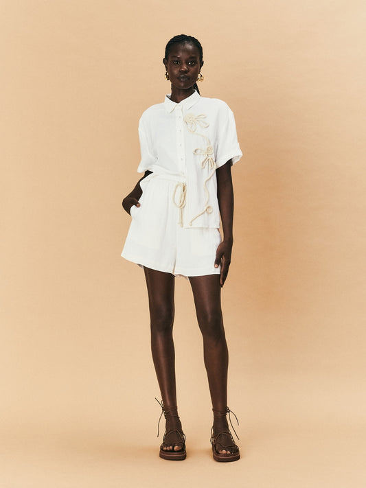 A young woman wearing a white La Maise Lillie short with gold accents on a beige background.