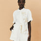 A young woman wearing a white La Maise Lillie shirt with white flower embroidery on a beige background.