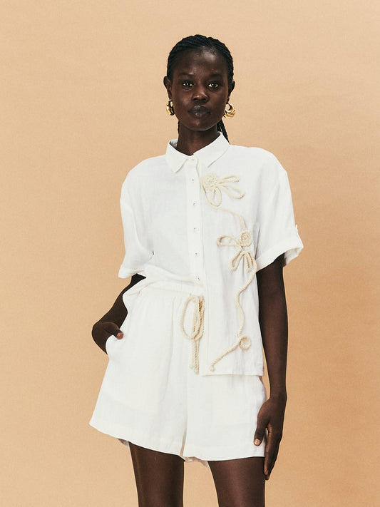 A young woman wearing a white La Maise Lillie shirt with white flower embroidery on a beige background.