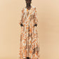 A young woman wearing a long, patterned La Maise Cannes Maxi Dress with seashell print against a beige background.