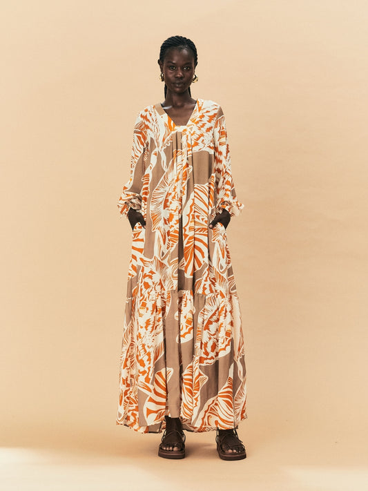 A young woman wearing a long, patterned La Maise Cannes Maxi Dress with seashell print against a beige background.