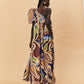 A woman wearing a one-shoulder, La Maise Dijon maxi dress on a beige background.