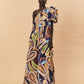 A woman wearing a one-shoulder, La Maise Dijon maxi dress on a beige background.