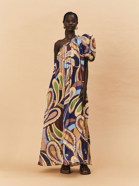 A woman wearing a one-shoulder, La Maise Dijon maxi dress on a beige background.