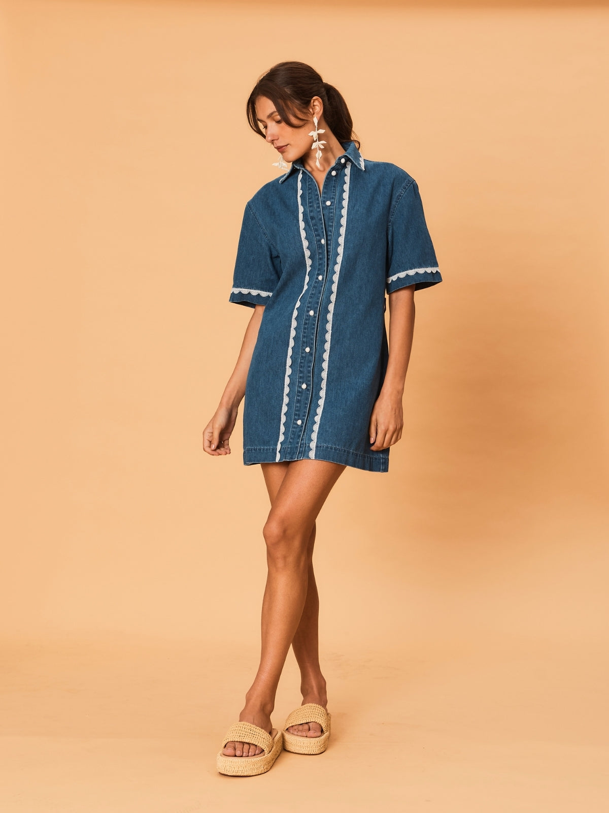 A young woman wearing a blue La Maise Ellie denim mini dress with white trim on a beige background.