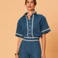 A young woman wearing La Maise Ellie denim shirt in blue denim with white lace trim on a beige background.