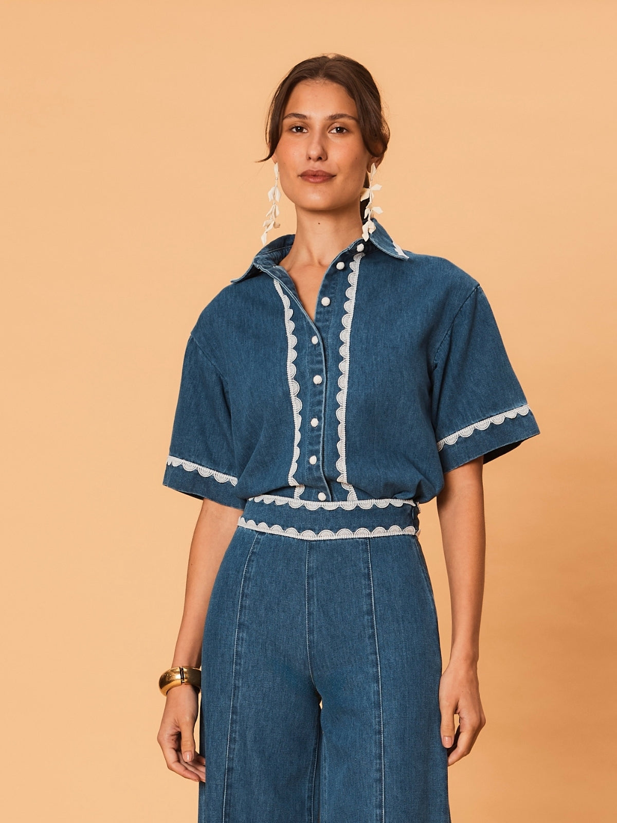 A young woman wearing La Maise Ellie denim shirt in blue denim with white lace trim on a beige background.