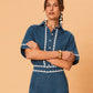 A young woman wearing La Maise Ellie denim shirt in blue denim with white lace trim on a beige background.