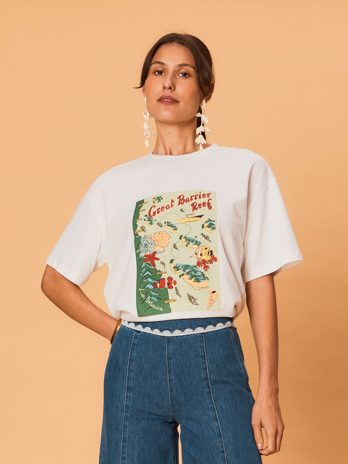 A young woman wearing a white La Maise Great Barrier Reef  tee on a beige background.