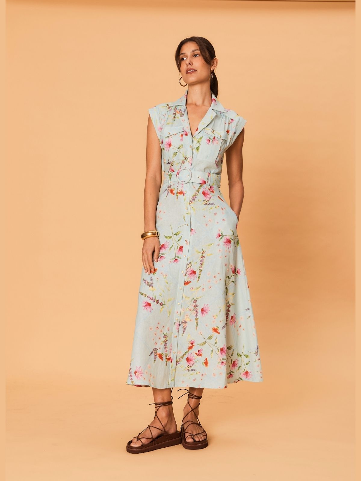 Model wearing a light blue midi dress with delicate floral print from La Maise, sleeveless design, button-through front and waist belt, styled with flat leather sandals.
