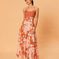 A young woman is wearing the Akira Maxi Dress by La Maise Studio, a sleeveless maxi dress with thin shoulder straps, a fitted striped bodice, and a flowing full-length skirt with a soft bird and floral print in warm rust and blush tones.