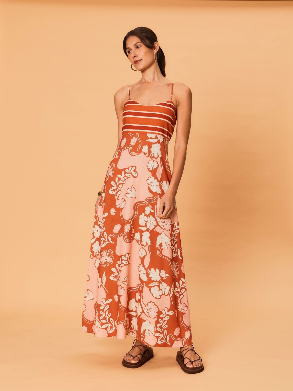 A young woman is wearing the Akira Maxi Dress by La Maise Studio, a sleeveless maxi dress with thin shoulder straps, a fitted striped bodice, and a flowing full-length skirt with a soft bird and floral print in warm rust and blush tones.