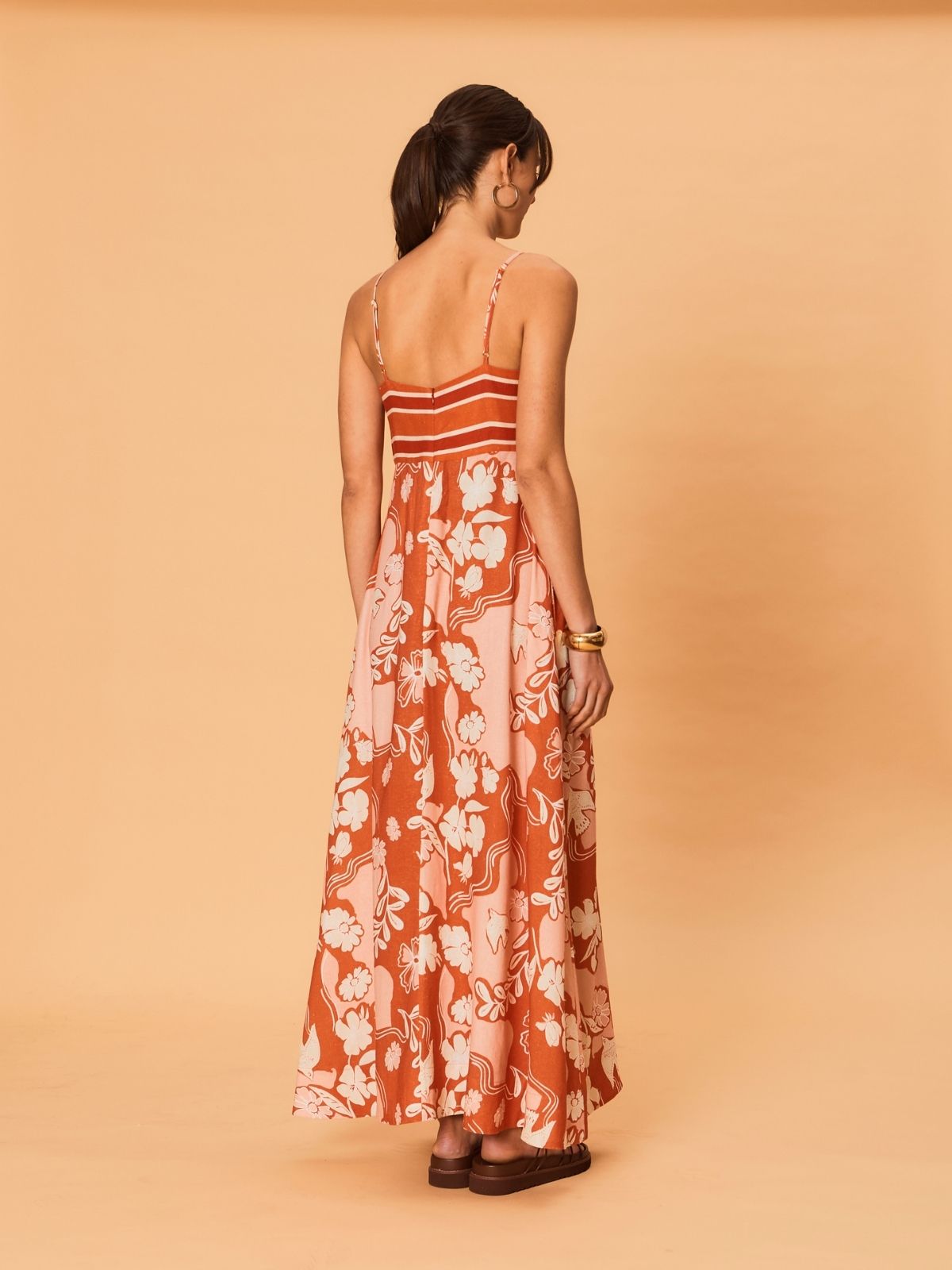 A young woman is wearing the Akira Maxi Dress by La Maise Studio, shown from the back with thin shoulder straps, a striped upper bodice, and a relaxed, flowing skirt featuring a bird and floral pattern that falls to the ankles.