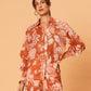 A young woman wearing the Akira Blouse by La Maise Studio, featuring a terracotta and blush bird and floral print, long sleeves and a button-front design, styled with matching printed shorts.