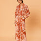 A young woman wearing the Akira Pant – Bird Floral by La Maise Studio, featuring wide-leg pants in a terracotta and blush floral print, styled with flat leather sandals.