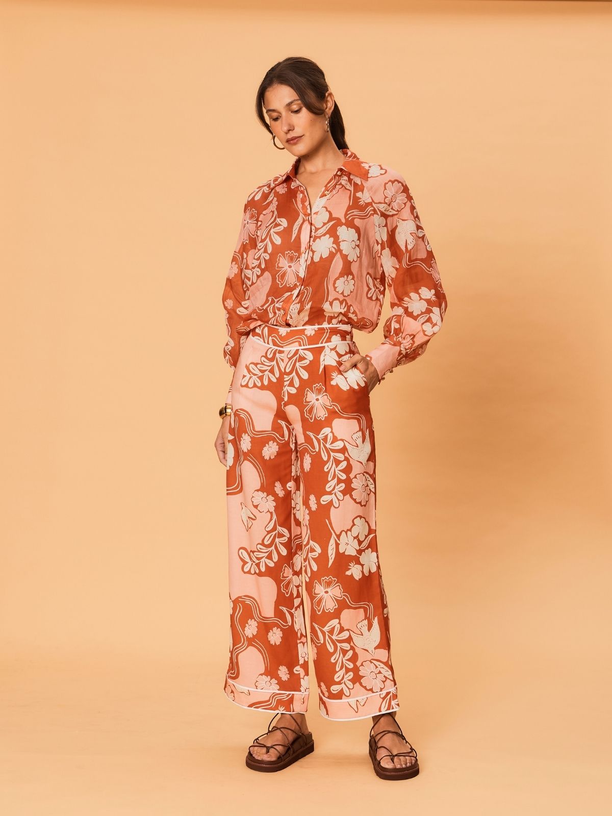 A young woman wearing the Akira Pant – Bird Floral by La Maise Studio, featuring wide-leg pants in a terracotta and blush floral print, styled with flat leather sandals.