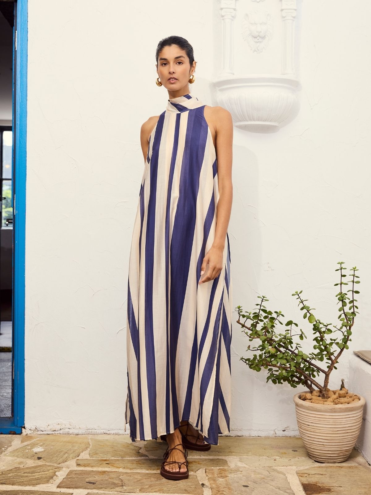 A young woman wearing the Mila Maxi Dress – Stripe by La Maise Studio, featuring a sleeveless high-neck design with vertical navy and cream stripes and a flowing maxi-length silhouette.