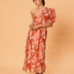 A young woman wearing the La Maise Studio Akira One Shoulder Dress in a bird floral print, featuring a one-shoulder neckline, soft puff sleeve, and flowing midi-length skirt in warm terracotta tones.
