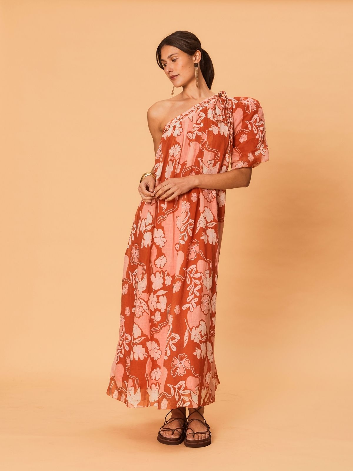 A young woman wearing the La Maise Studio Akira One Shoulder Dress in a bird floral print, featuring a one-shoulder neckline, soft puff sleeve, and flowing midi-length skirt in warm terracotta tones.