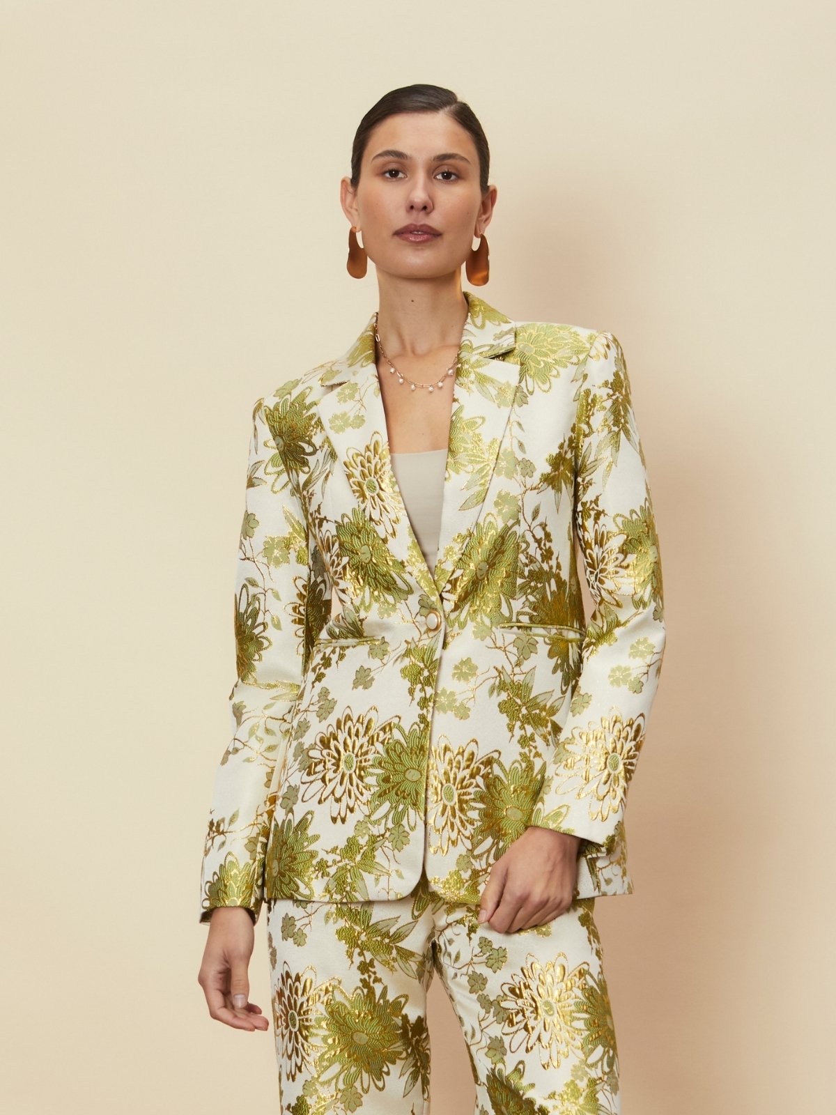 A model wearing the Dahlia Blazer by La Maise Studio, a tailored floral jacquard blazer in soft cream with green and gold botanical detailing, featuring a structured lapel collar, single-button closure and long sleeves, styled with the matching Dahlia Skirt against a neutral studio background.