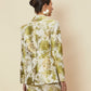 A rear view of a model wearing the Dahlia Blazer by La Maise Studio, showcasing the tailored back shape, structured shoulders and detailed floral jacquard fabric in cream, green and gold tones, styled as a matching set in a studio setting.