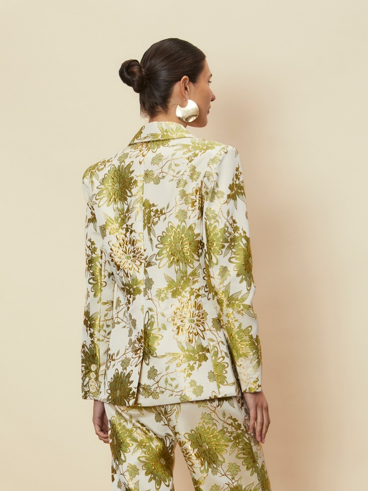 A rear view of a model wearing the Dahlia Blazer by La Maise Studio, showcasing the tailored back shape, structured shoulders and detailed floral jacquard fabric in cream, green and gold tones, styled as a matching set in a studio setting.