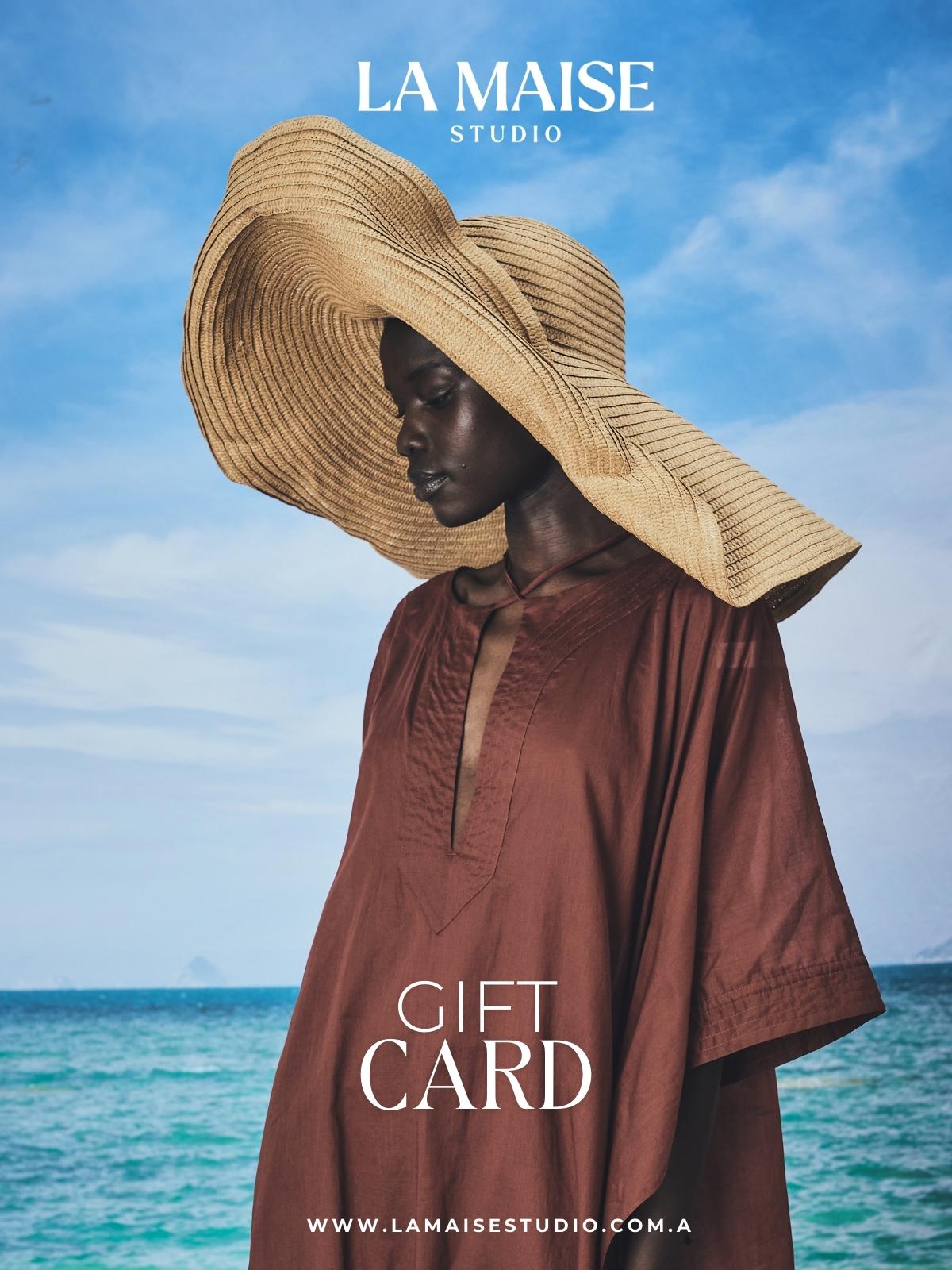 Person wearing a large straw hat and brown garment with 'La Maise Studio' branding on a beach.